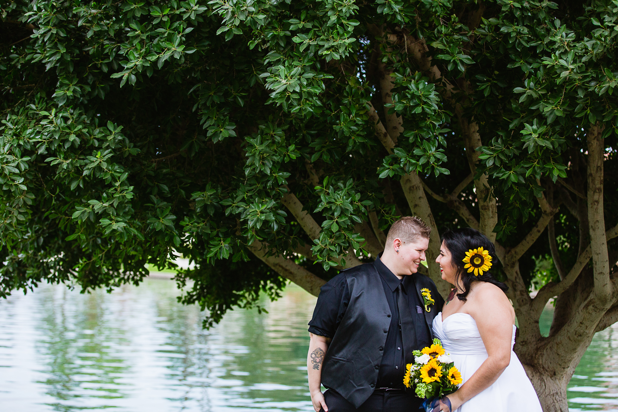 Same sex couple looking at each other in front of a tree and lake by Phoenix wedding photographer PMA Photography.