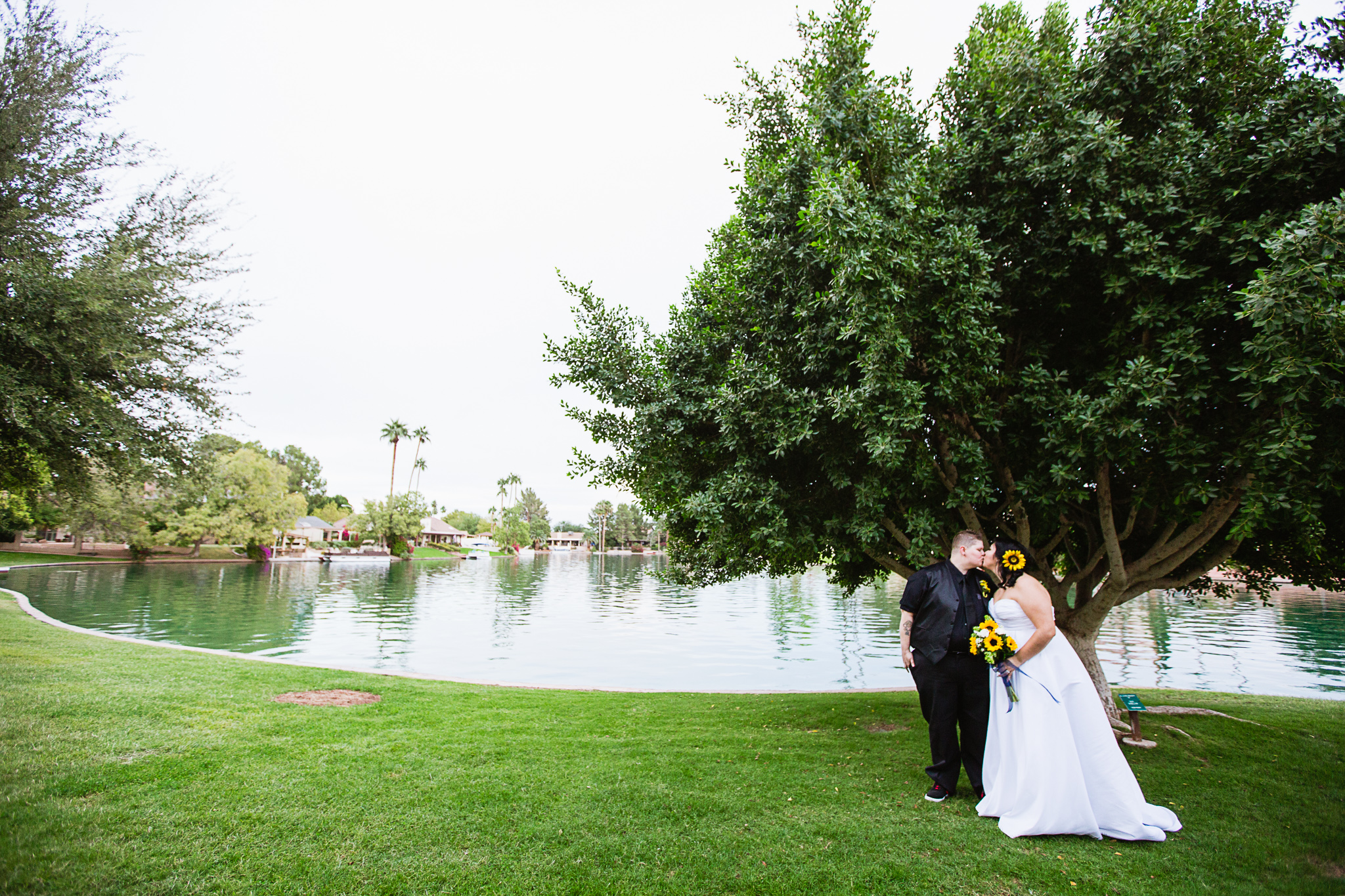 Same sex couple kissing in front of a tree and small lake in a park on their wedding day by Phoenix wedding photographers PMA Photography.
