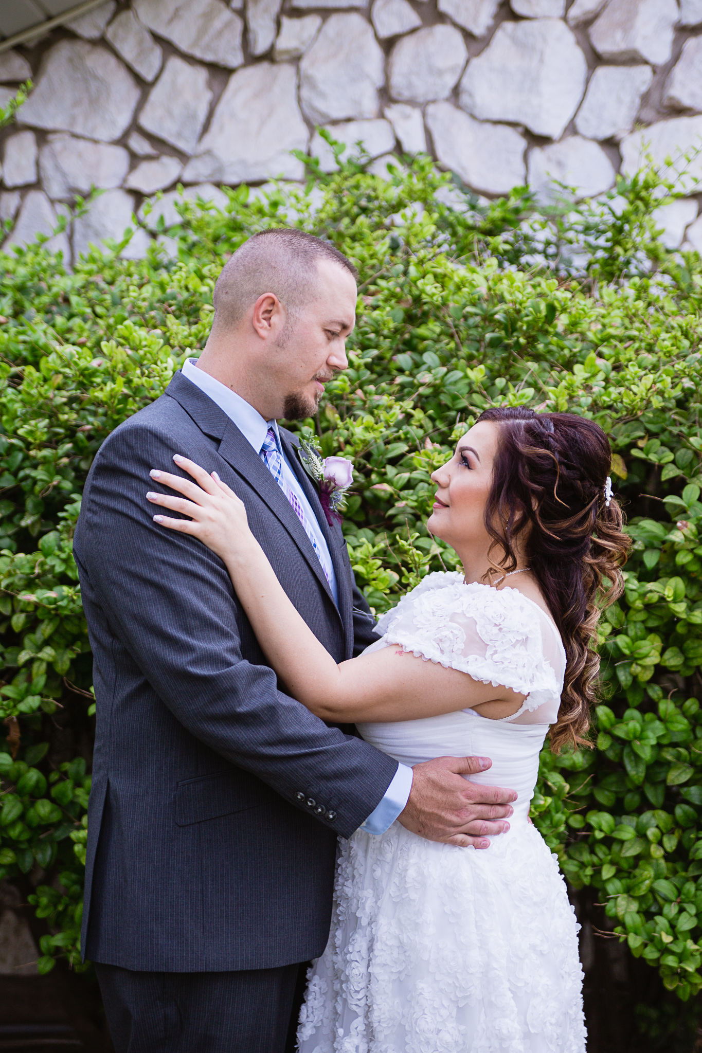Bride and groom look at each other in front of a garden bush by PMA Photography.