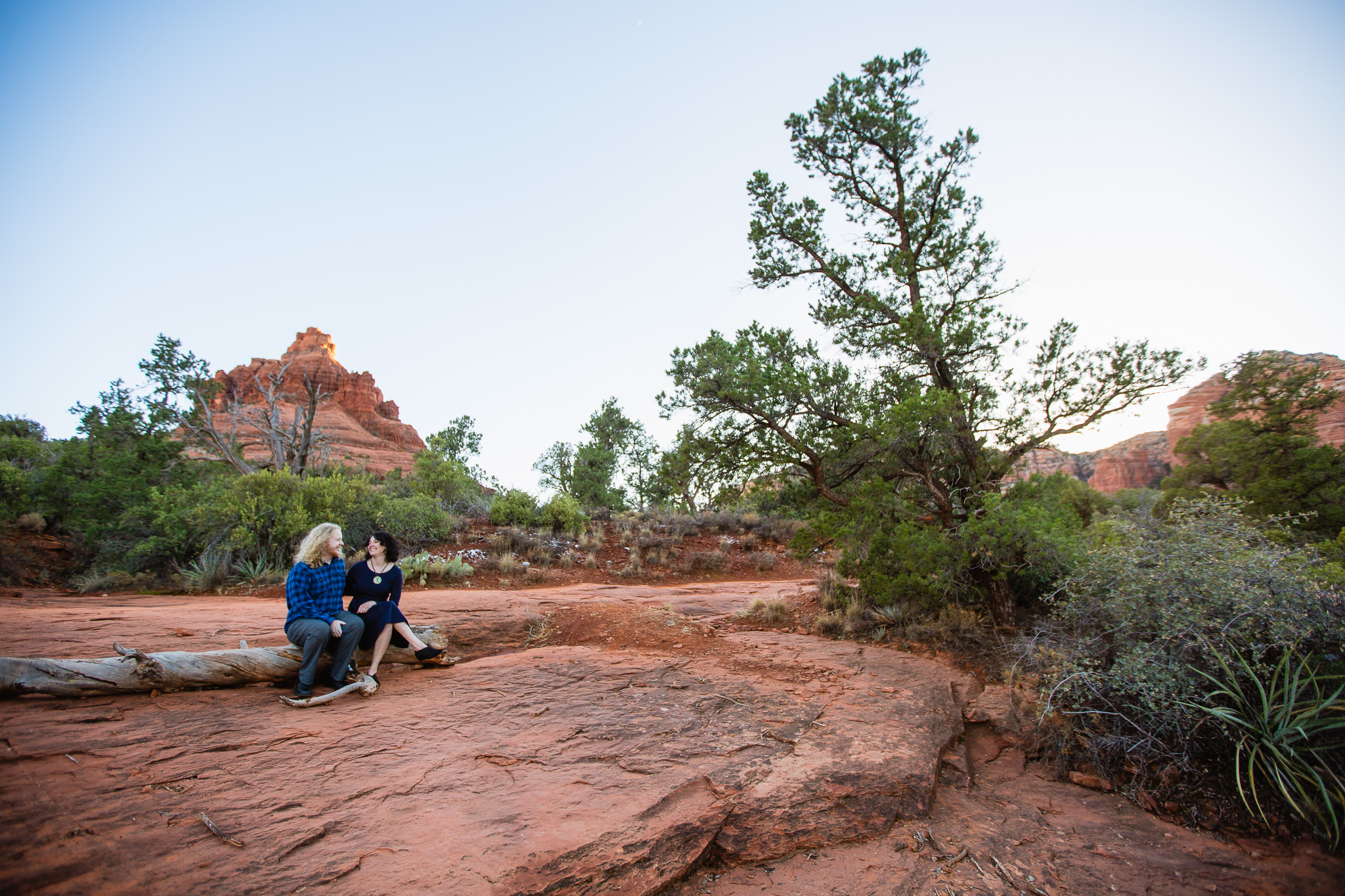 Outdoorsy couple laughing while sitting on a log during an engagement session at Bell Rock by PMA Photography.