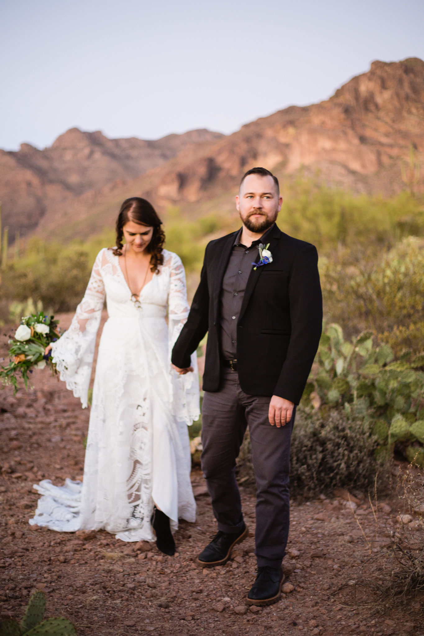 Witchy bohemian black white and gold inspired groom leading bride down trail at a Superstition Mountains styled wedding surrounded by the beauty of the desert.