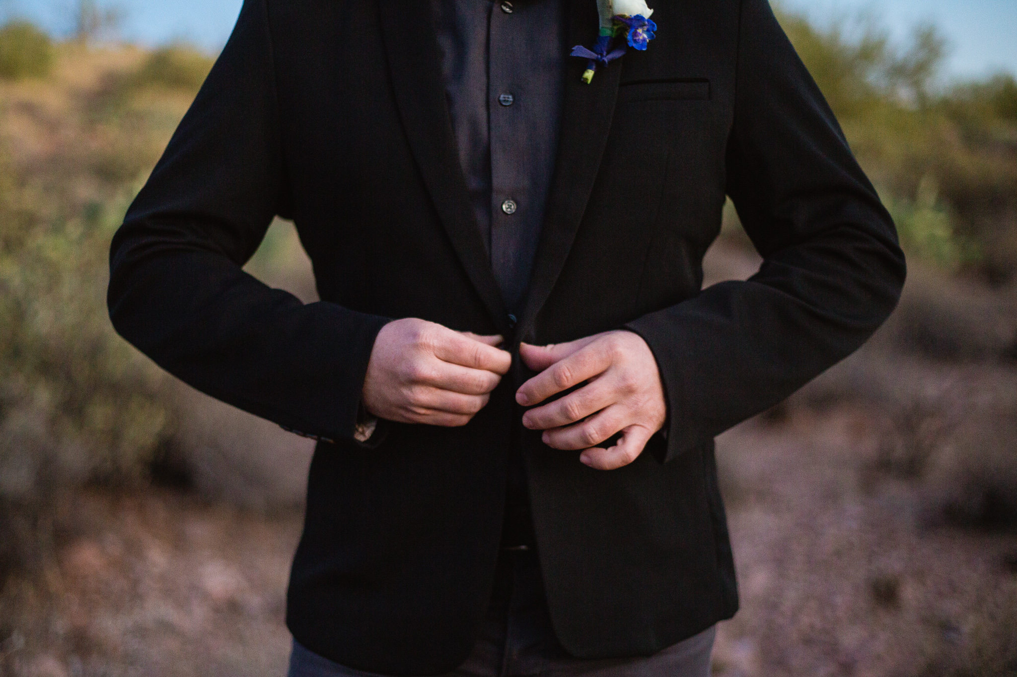Detail photograph of groom buttoning his jacked on his wedding day in his black and dark grey casual suit.