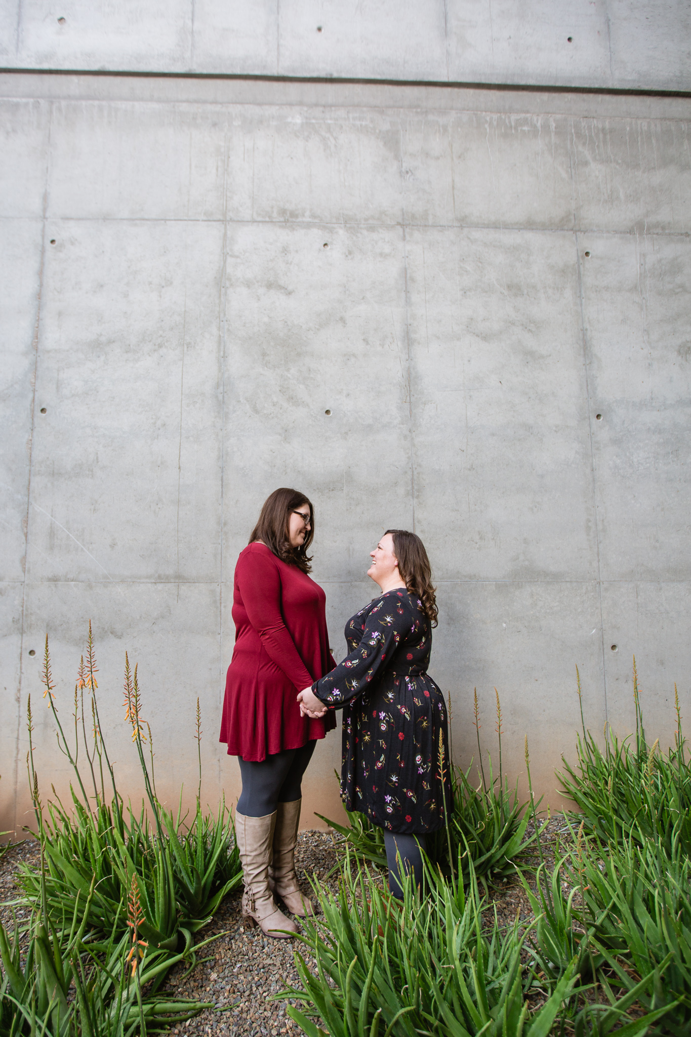 LGBTQ couple in burgundy, tan, and black coordinating outfits pose in front of industrial wall downtown for their engagement session. 