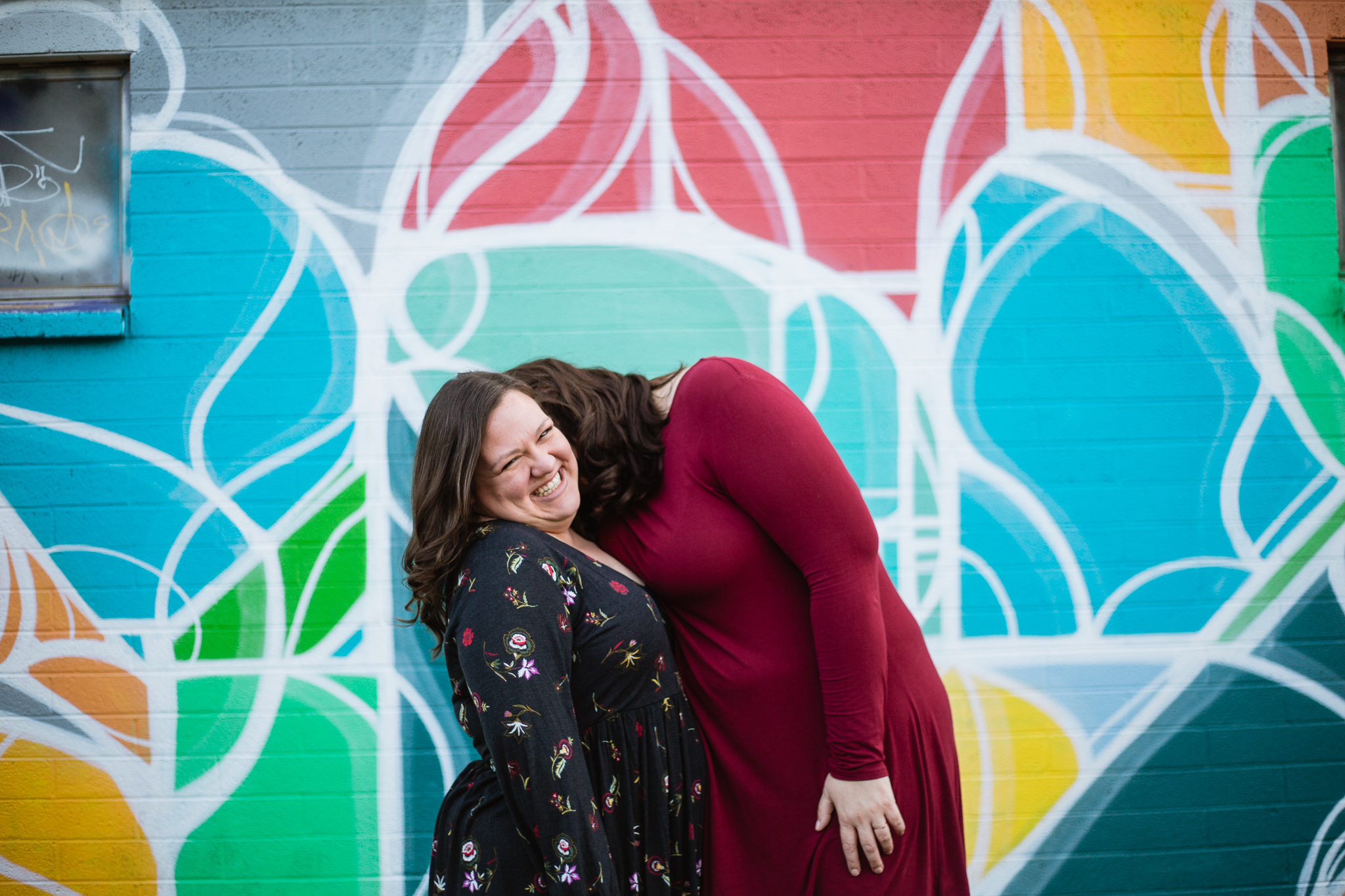 LGBT couple laughing together in front of a mural for their engagement photography session in downtown Phoenix at Roosevelt Row.