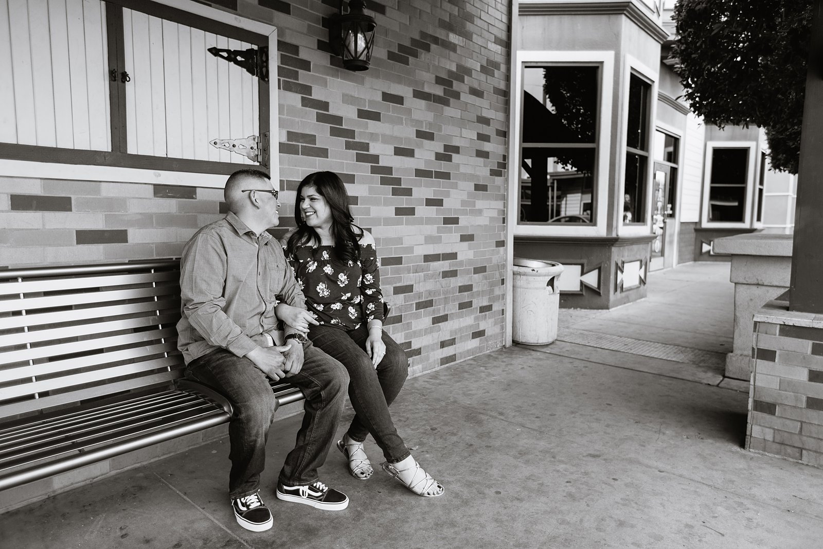 Black and white image of couple sitting on bench in Gilbert heritage district at their engagement session by wedding photographers PMA Photography.
