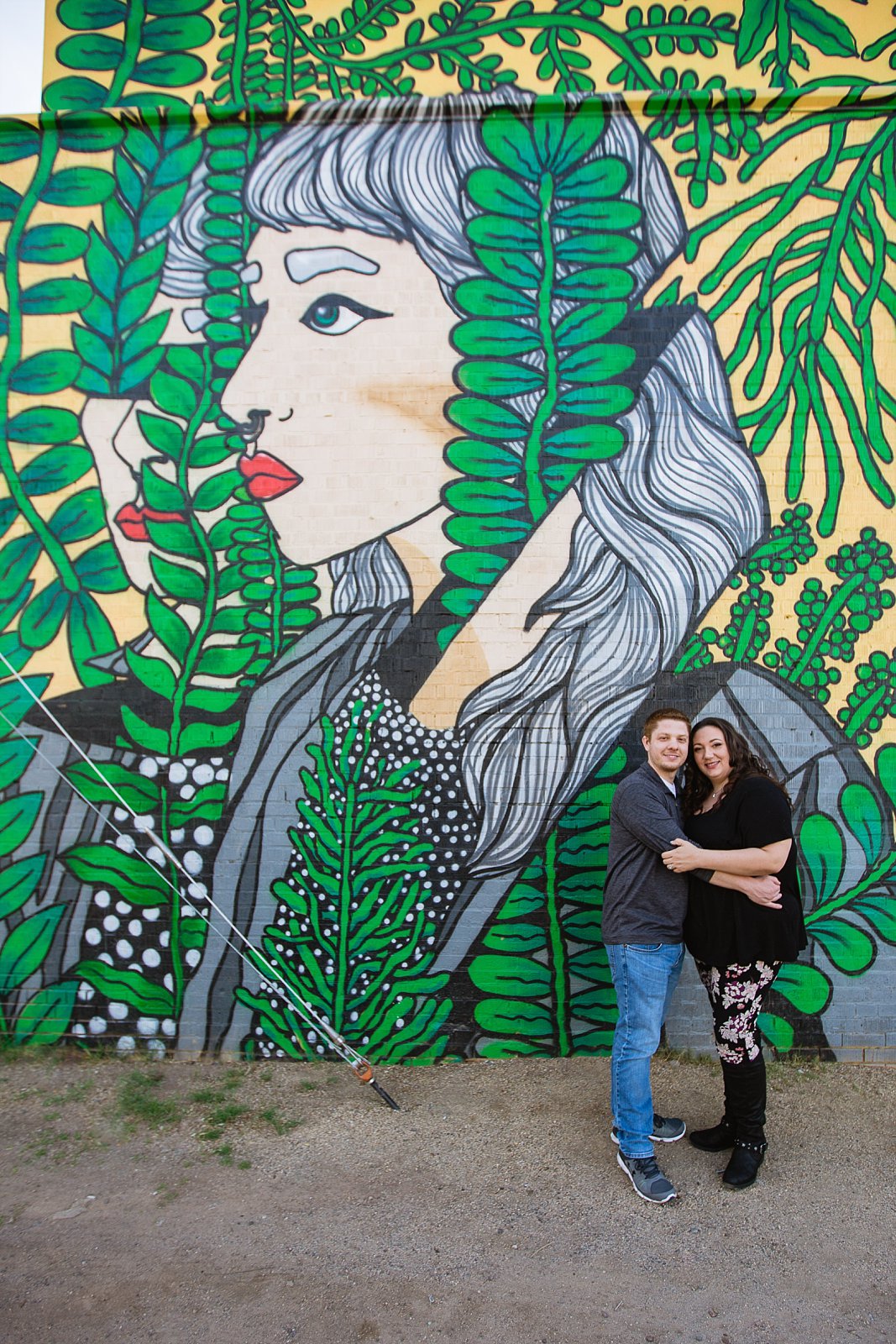 Couple pose in front of art mural of girl with green plants for their Roosevelt Row Art District engagement session by PMA Photography.