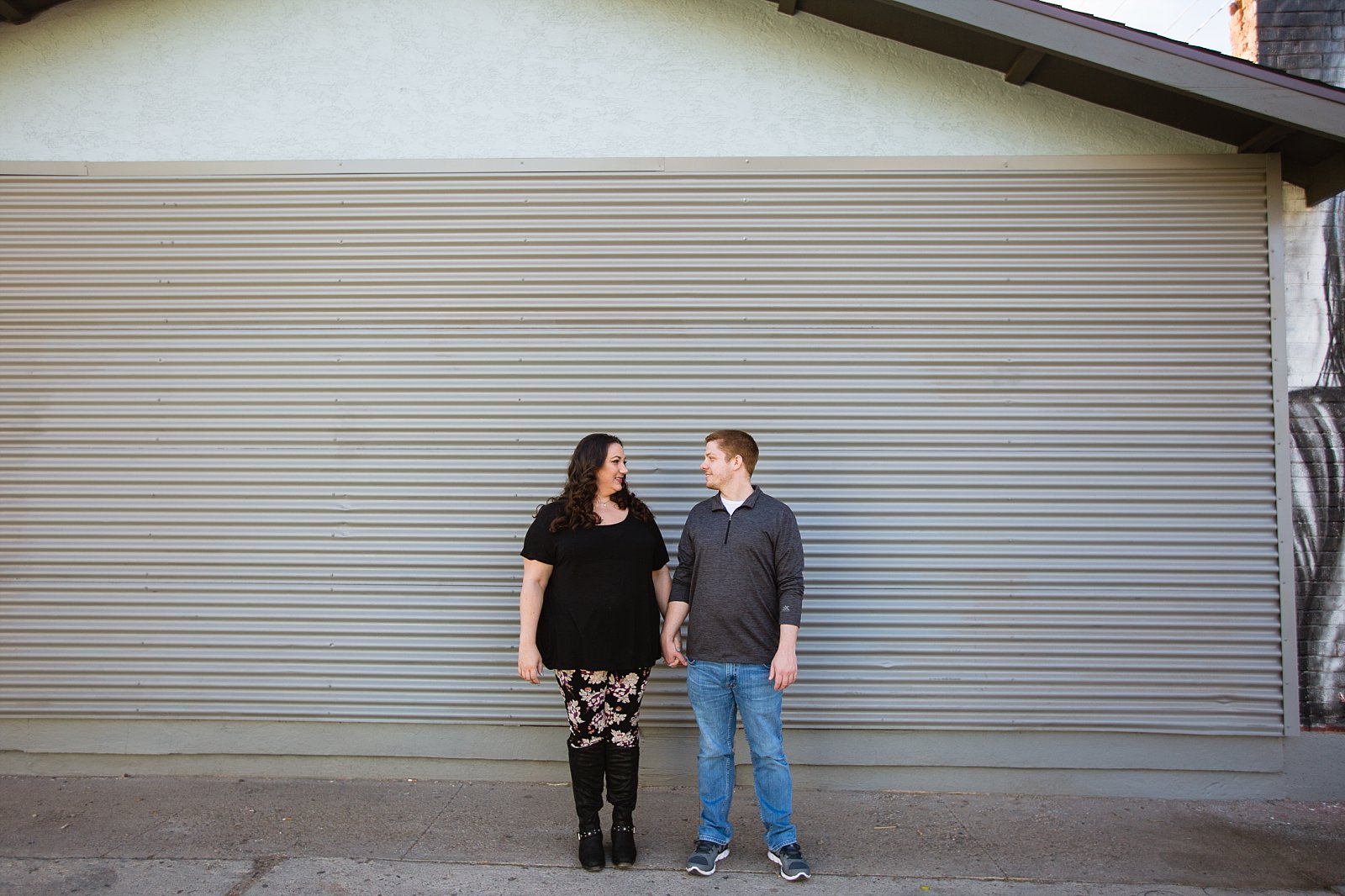 Couple stand holding hands looking at each other in front of grey siding for their downtown Phoenix engagement session by photographer PMA Photography.