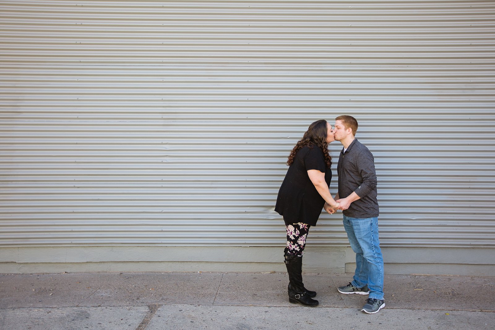 Couple stand holding hands kissing in front of grey siding for their downtown Phoenix engagement session by photographer PMA Photography.