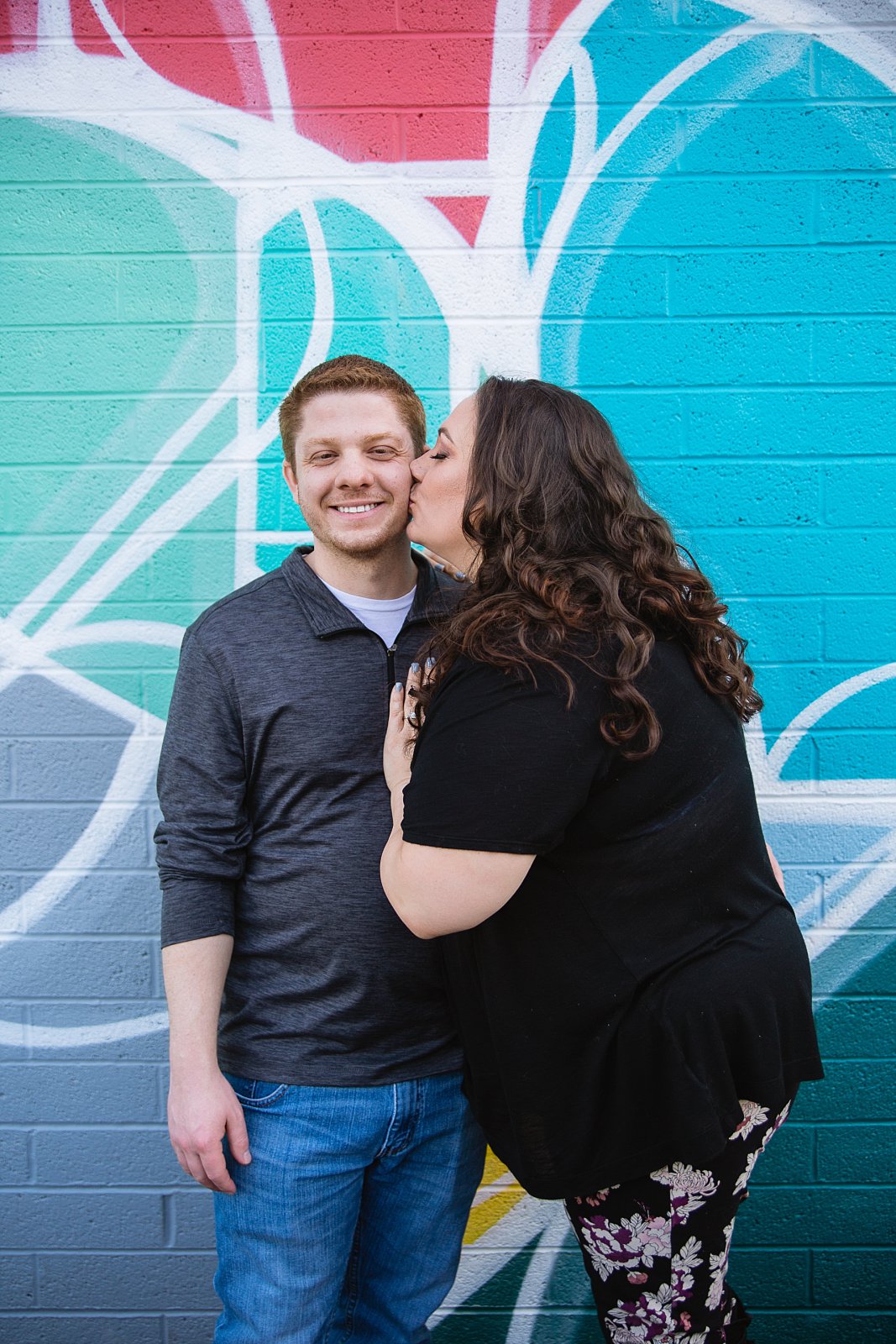 Future bride kissing fiance's check in front of art mural during their Roosevelt Row Art District engagement session by PMA Photography.