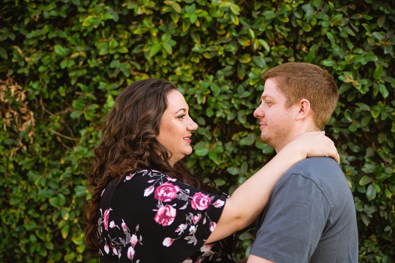 Couple looking at each other in front of living plant wall during their downtown Phoenix engagement session by PMA Photography.