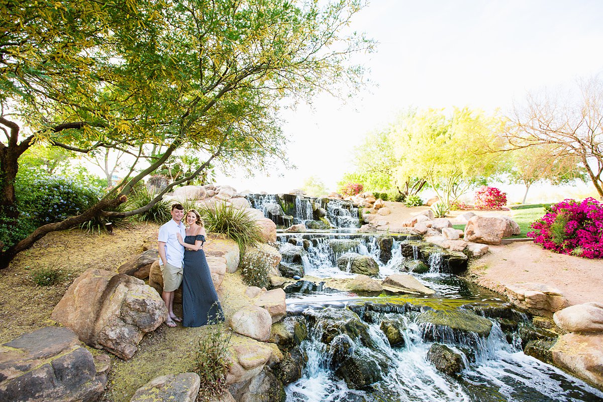 Couple posing during their engagement session in front of blossoming Palo Verde and waterfall at Anthem Community Park by PMA Photography. 