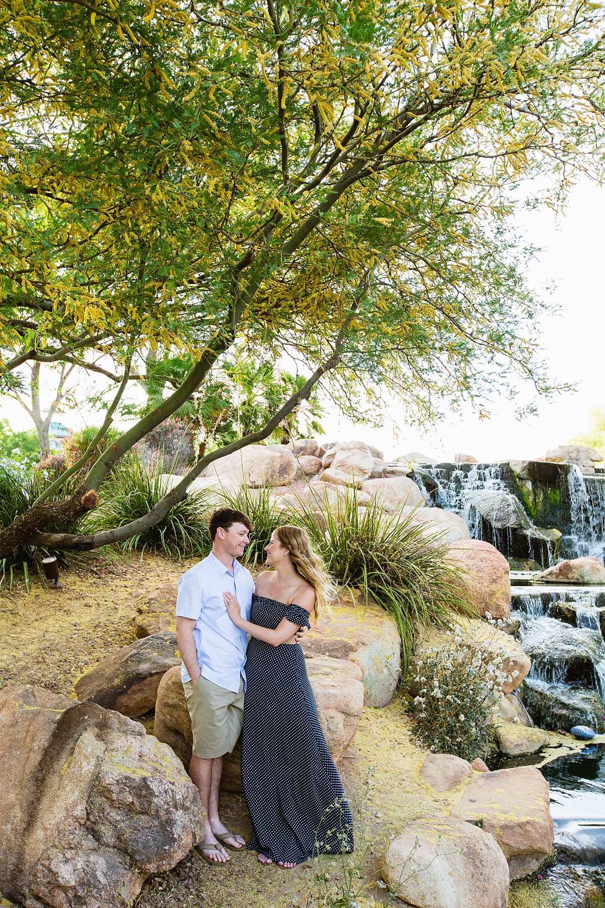 Couple posing during their engagement session in front of blossoming Palo Verde and waterfall at Anthem Community Park by PMA Photography. 