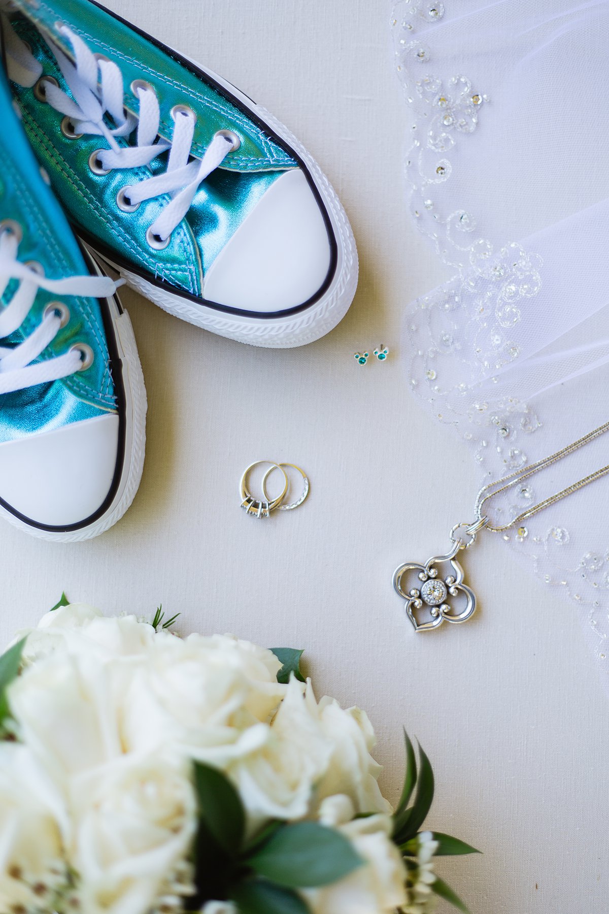 Bride's wedding details of metallic Turquoise converse, white roses, blue Mickey ear rings, wedding bands, necklace, and veil by PMA Photography.