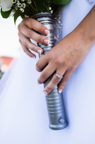 Close up image of bride's wedding bands holding her bouquet in a Star Wars light saber hilt by PMA Photography.