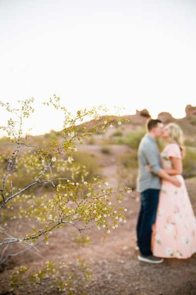 Couple sharing a kiss behind a desert bush during their summer engagement session at Papago Park by PMA Photography.