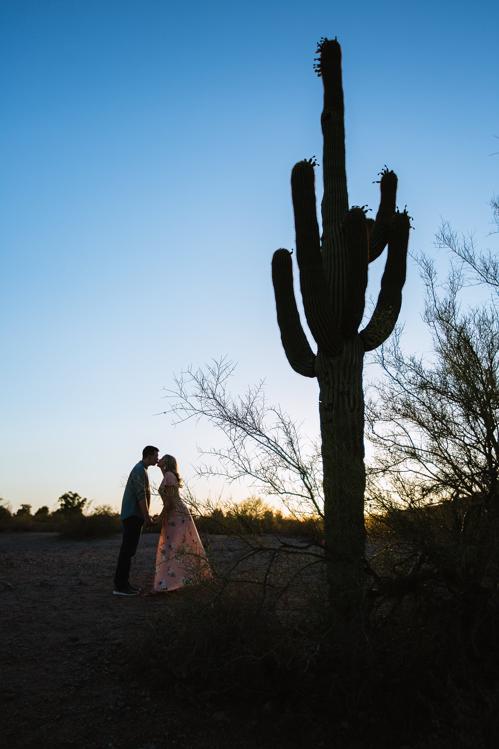 Couple share a kiss next to a silhouette of a saguaro cactus as the sun sets by Arizona engagement photographer PMA Photography.