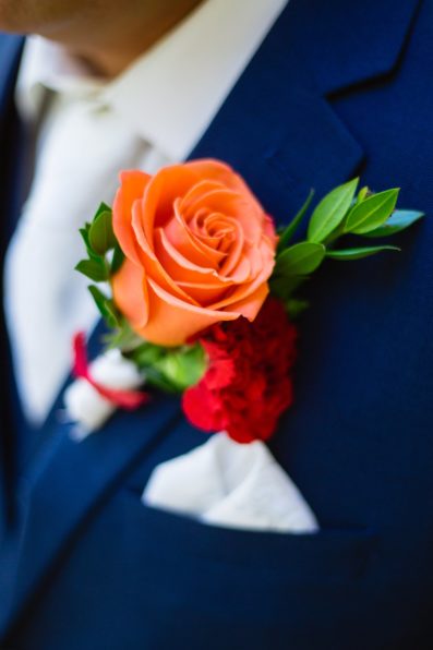 Groom's colorful orange and red rose boutonniere on his navy suit by PMA Photography.