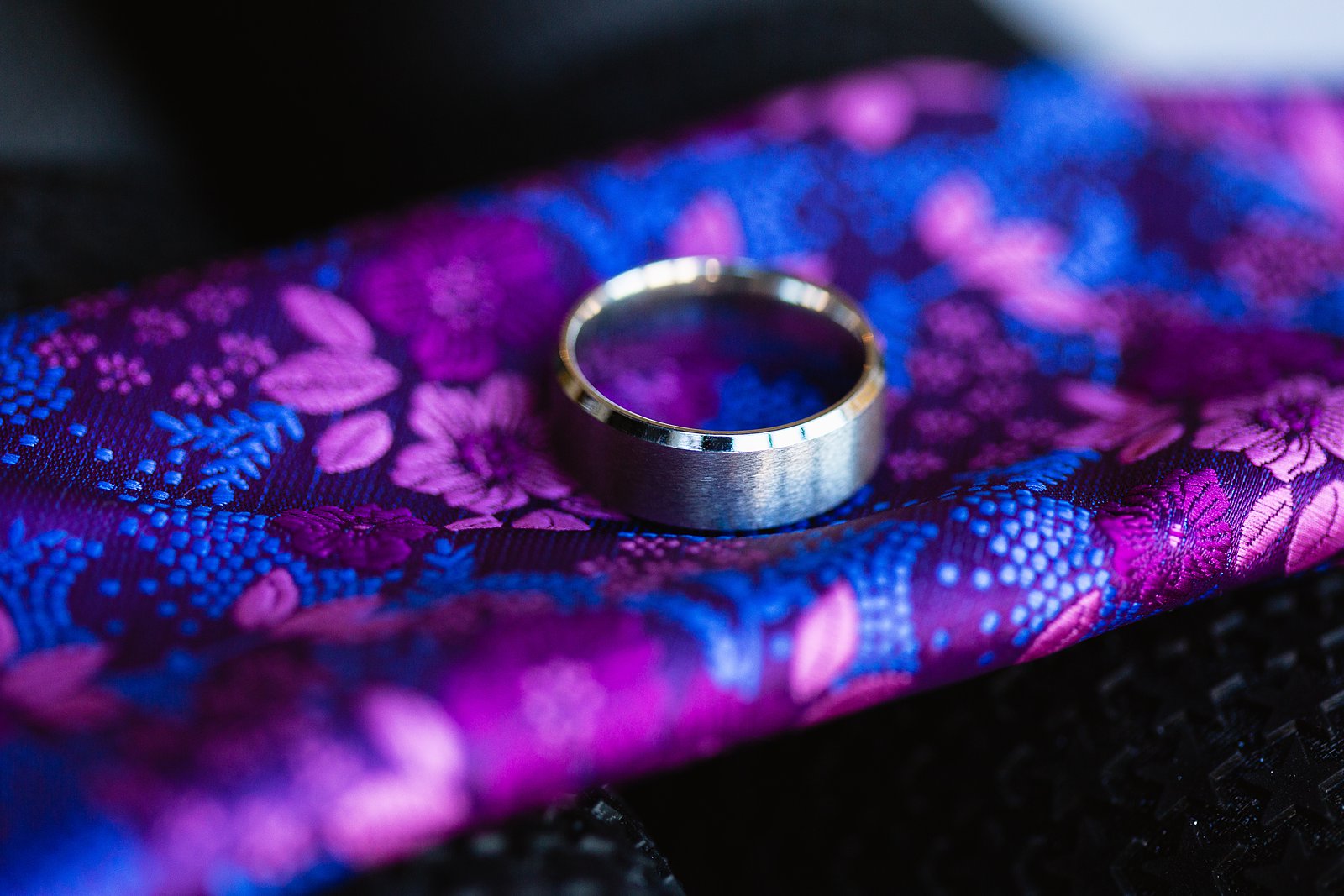 Simple silver wedding band on purple floral pocket square by PMA Photography.