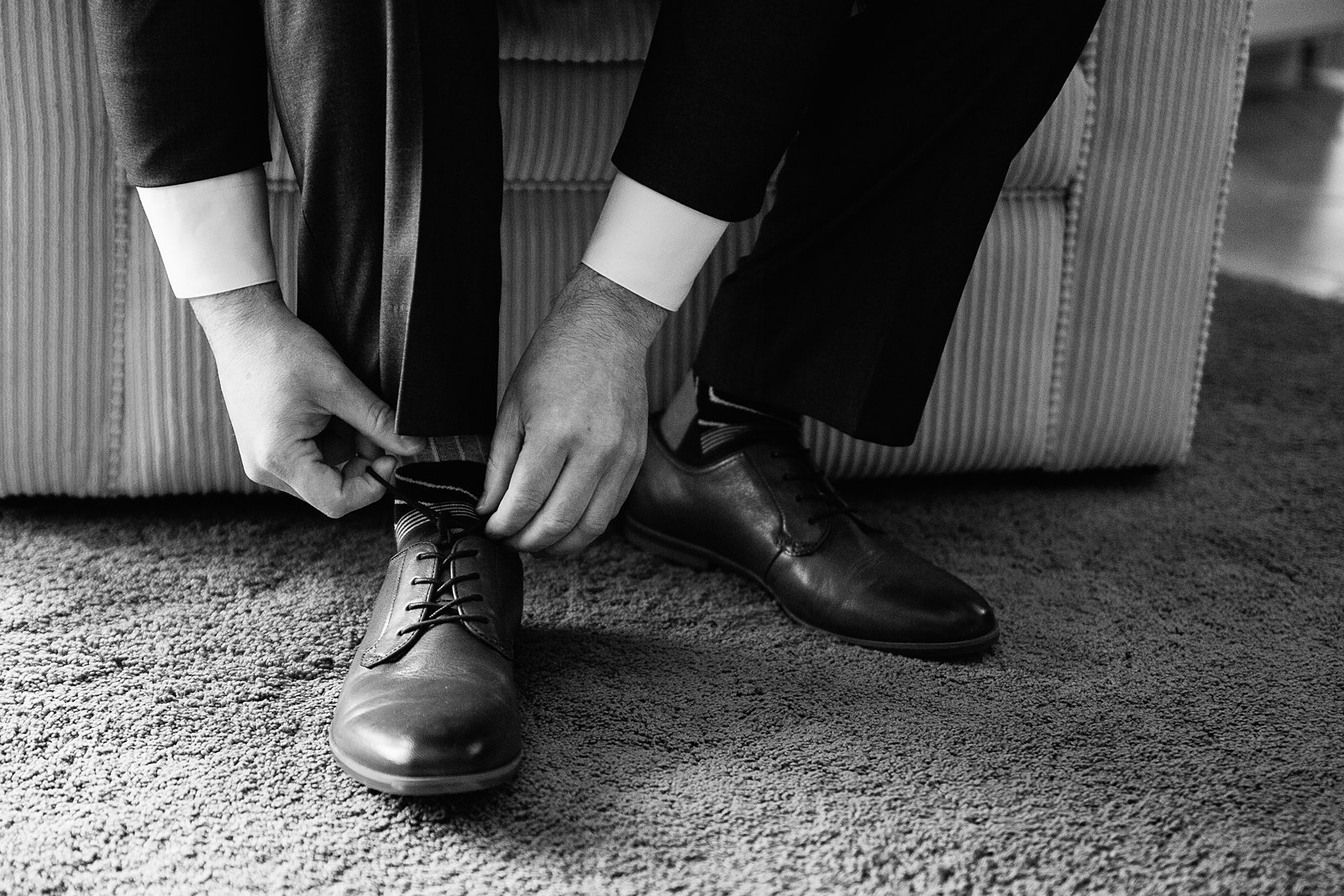 Black and white image of groom tying his shoes while getting ready for his wedding day by PMA Photography.