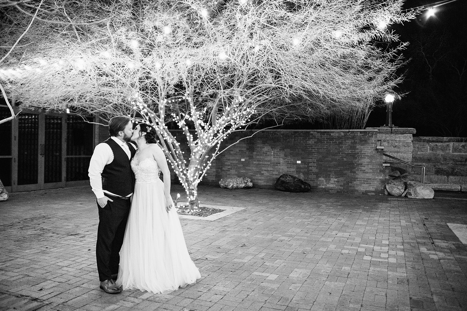 Black and white image of bride and groom kissing in front of a tree decorated in lights by Arizona wedding photographer PMA Photography.
