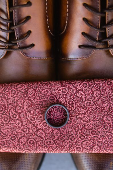 Groom's wedding day details of a black wedding band on a red copper tie and tan shoes by PMA Photography.