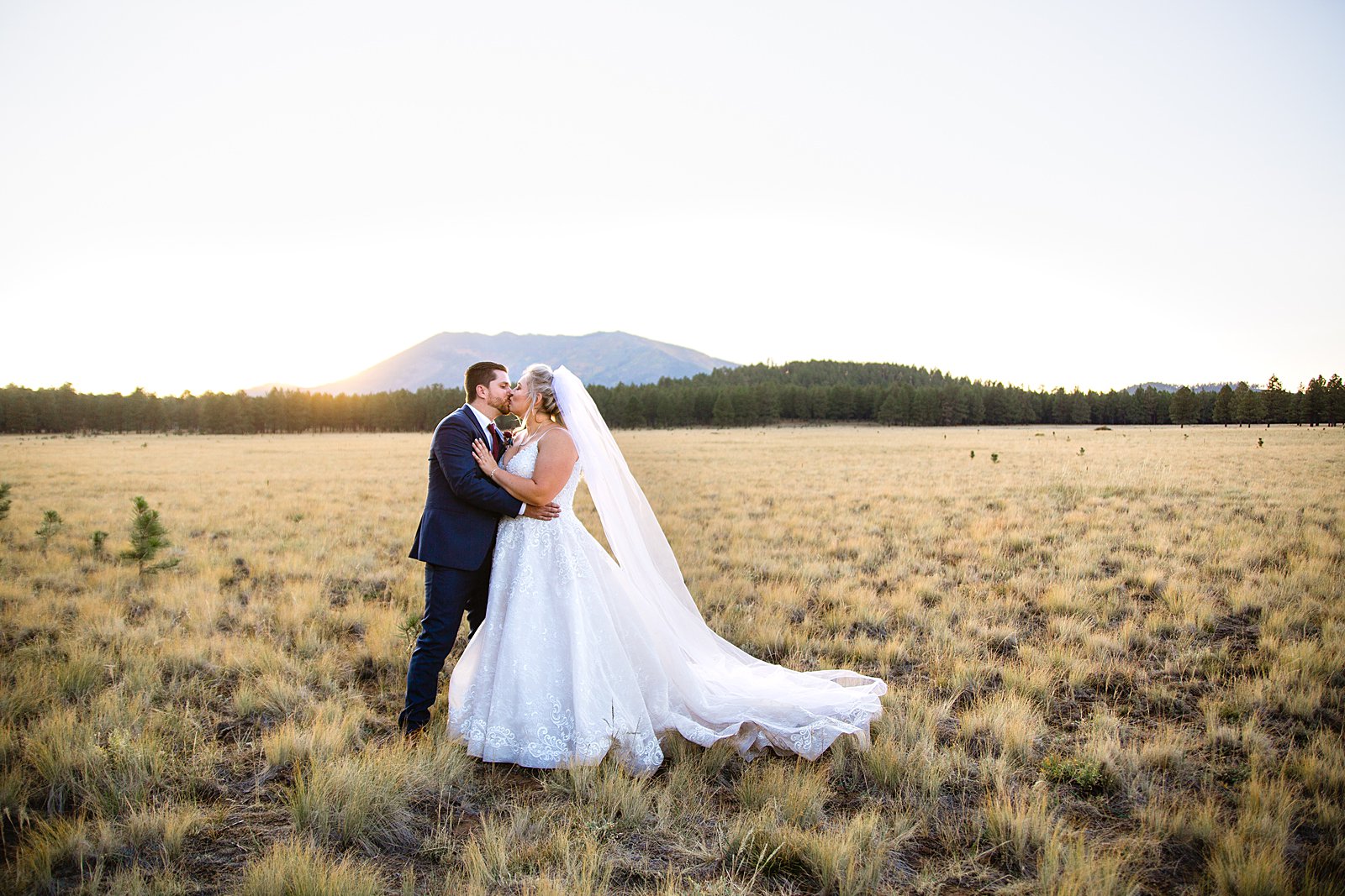 Bride and groom share a kiss in a field with a mountain backdrop at sunset by Flagstaff wedding photographers PMA Photography.