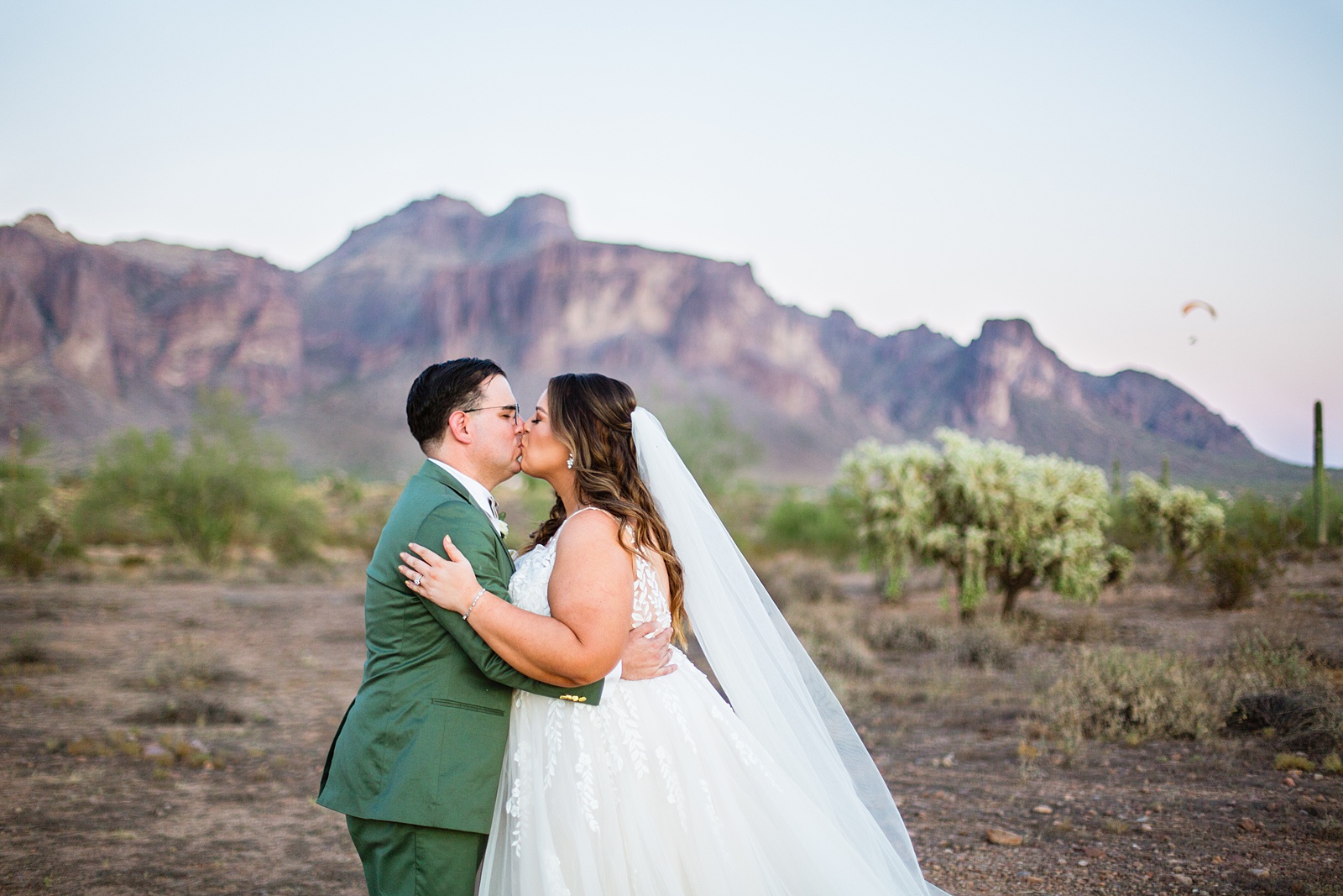 Bride & Groom shares a kiss as parasailer flies in the desert behind them during their a Phoenix desert intimate wedding by Phoenix wedding photographer Juniper and Co Photography.
