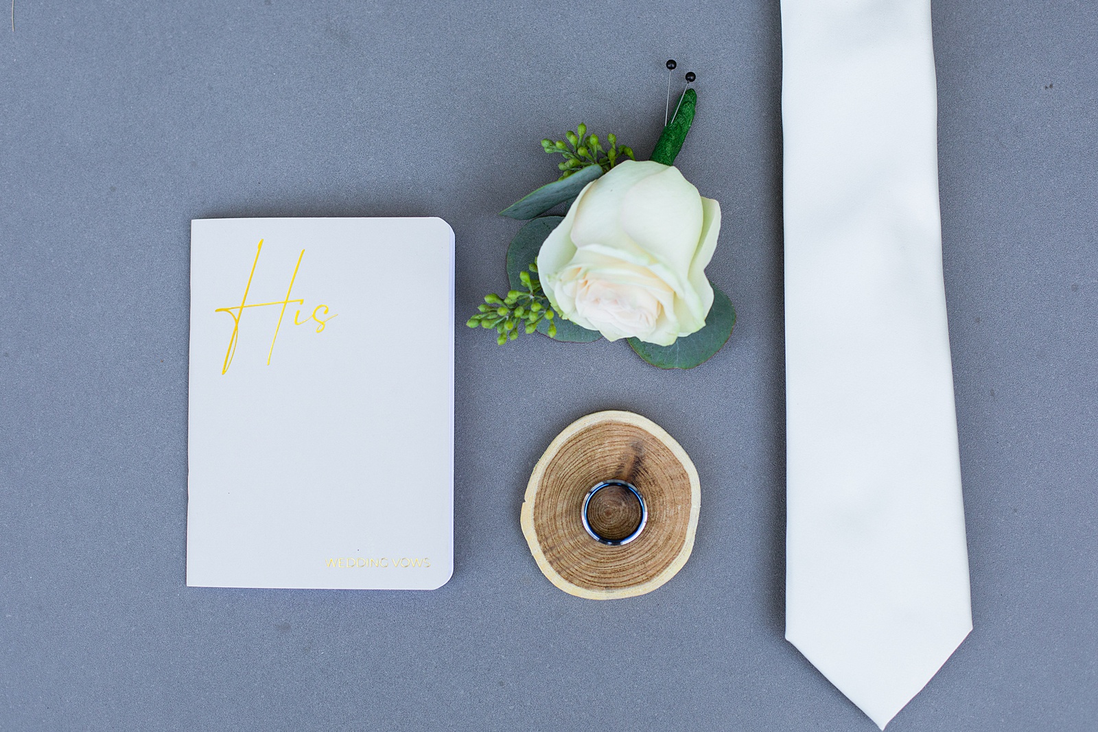Groom's wedding day details of vow book, boutonniere, white tie and wedding band by Juniper and Co Photography.